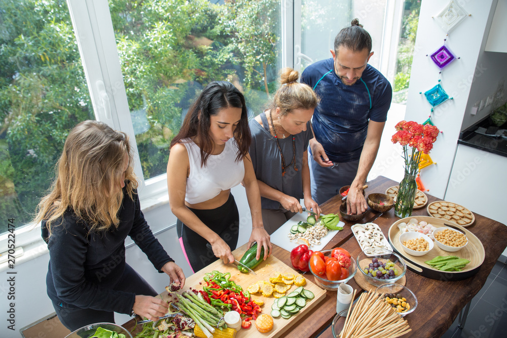 Naklejka premium Serious people cooking vegetables at kitchen table. Man and women standing at table with fresh vegetables and window with green view in background. Healthy cooking concept.