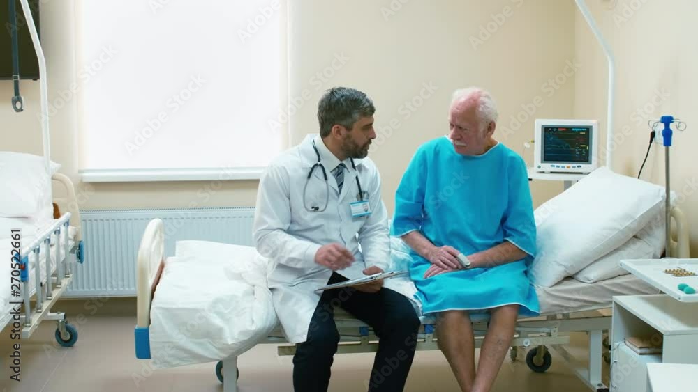 Old man sitting on therapy bad in hospital attire and explaining his male doctor, who sitting near, his symptoms Old man sitting on therapy bad in hospital attire and explaining his male doctor, who sitting near, his symptoms
