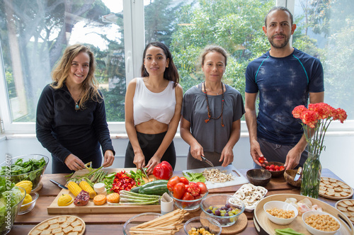 Smiling people posing at camera and cooking in kitchen. Man and women standing at table with fresh vegetables and window in background. Healthy cooking and food concept. Front view.