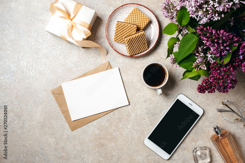 Minimal home office desk workspace with white paper card, kraft envelope, coffee cup, cookies, lilac flowers bouquet, perfume bottle, glasses, smartphone. Flat lay, top view social media background.