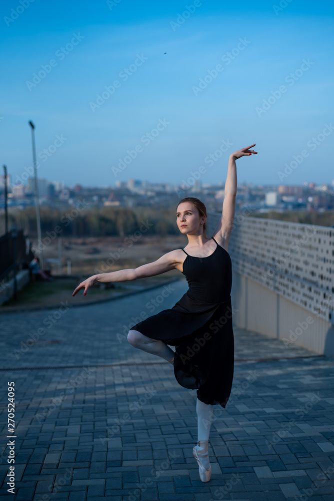 Ballerina in a tutu posing next to the fence. Beautiful young woman in black dress and pointe dancing over city background. Portrait of a gorgeous ballerina performing a dance outdoors