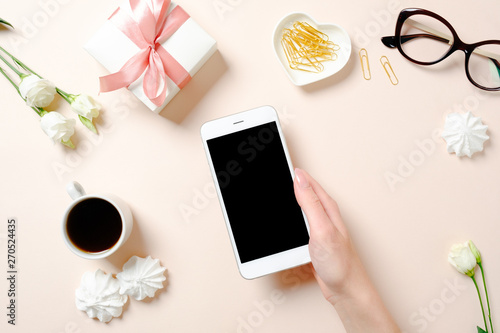 Flat lay home office desk. Female workspace with human hand holding smartphone, coffee cup, glasses, flowers, golden accessories on pink background. Top view feminine background.