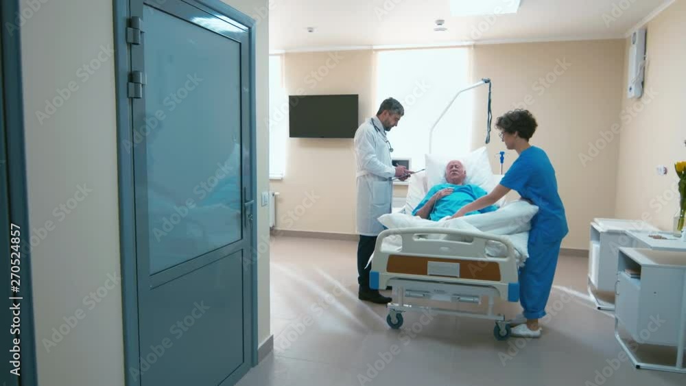 Hospital room where old man laying on bed while nurse covering him with blanket and doctor standing near and answering about symptoms Hospital room where old man laying on bed while nurse covering him with blanket and doctor standing near and answering about symptoms