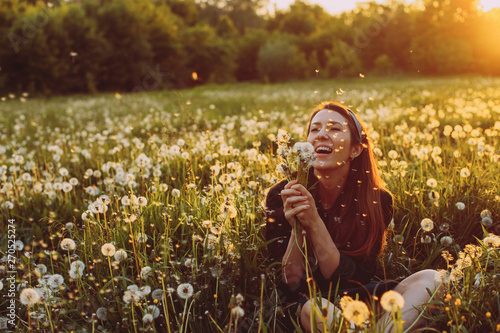Beautiful Young Woman lying on the field in green grass and blowing dandelion. Outdoors. Enjoy Nature. Healthy Smiling. Allergy free concept. Freedom