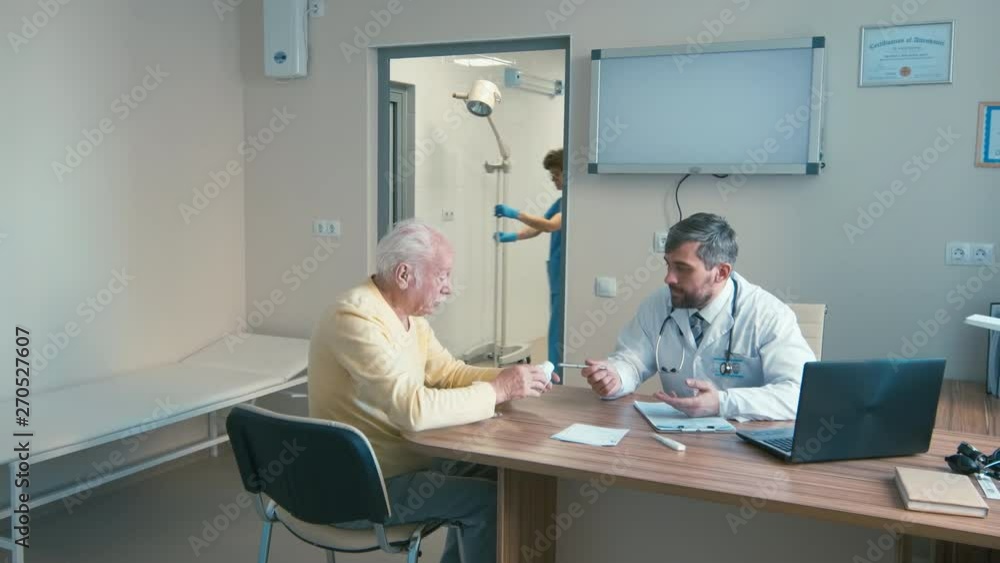 Old patient sitting in doctor's room and trying to read prescribing information to medicine, discussing it with doctor while nurse passing by with drop bottle in background Old patient sitting in doctor's room and trying to read prescribing information to medicine, discussing it with doctor while nurse passing by with drop bottle in background