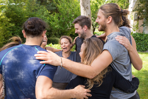 Group of happy people embracing each other after workout. Men and women in fitness apparels forming circle and hugging in park. Fitness and teambuilding concept photo