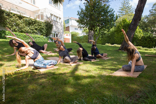 Peaceful people doing yoga on grass. Men and women sitting on mats and practicing asana, apartment building or hotel in background. Yoga club concept