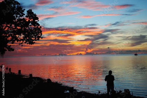 An unrecognizable man stands on the beach facing a blood-red sunset at the Saipan lagoon © raksyBH