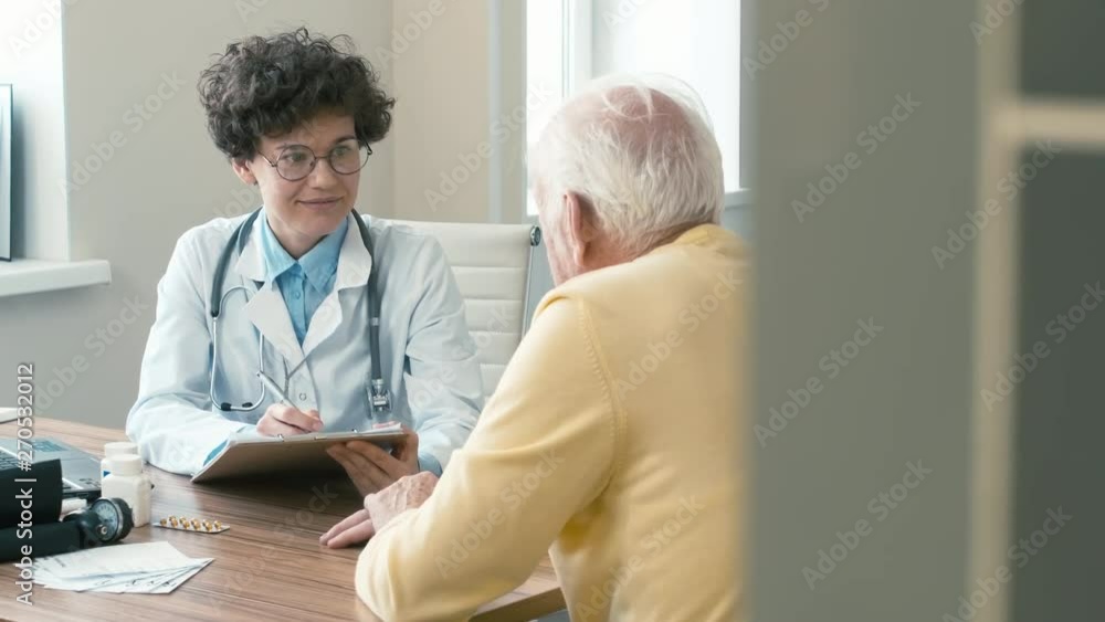 female doctor listening to complaints of elderly man who sitting opposite in consulting room, nodding head and doing notes