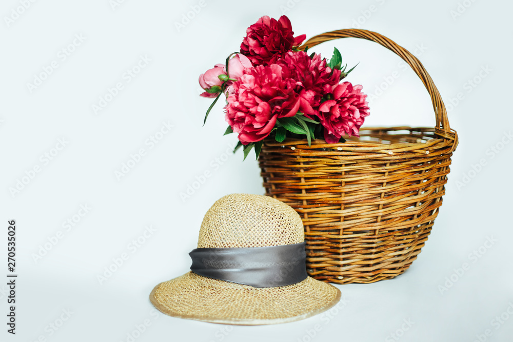 Straw hat with a bouquet of luxurious, lush coral and pink peonies. Flowers in a wicker basket on a white background. Straw hat with a bouquet of luxurious, lush coral and pink peonies. Flowers in a wicker basket on a white background.