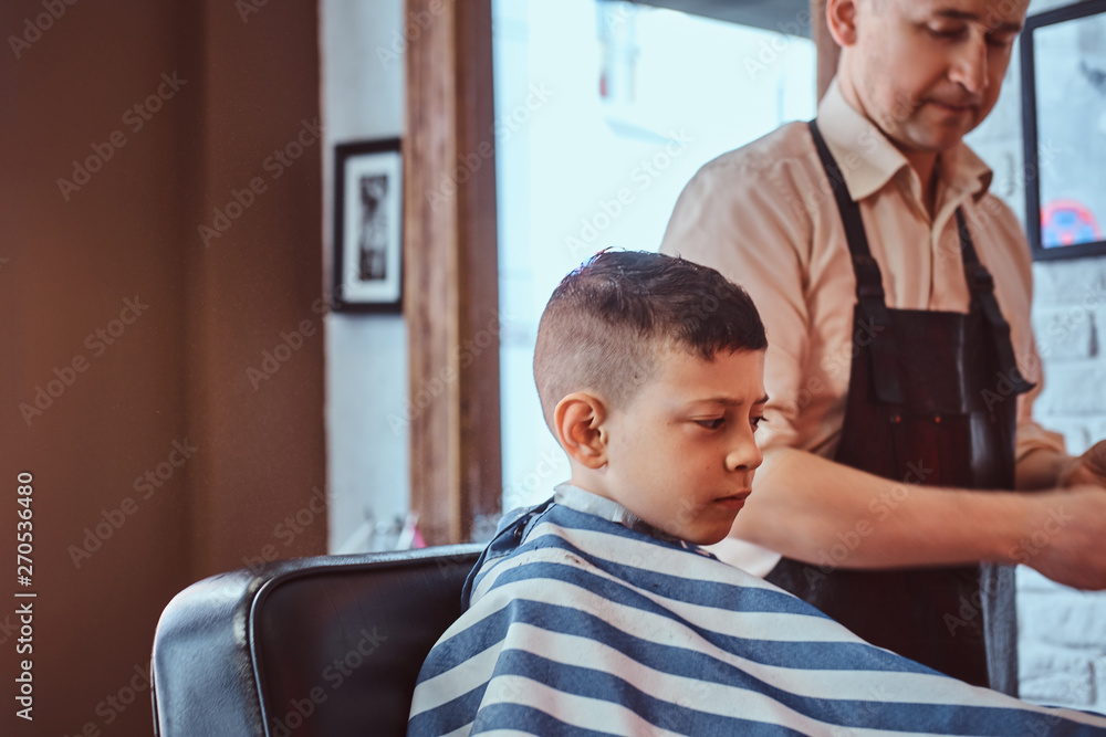 Obraz premium Attractive school boy is getting trendy haircut from mature hairdresser at fashionable hairdressing salon.
