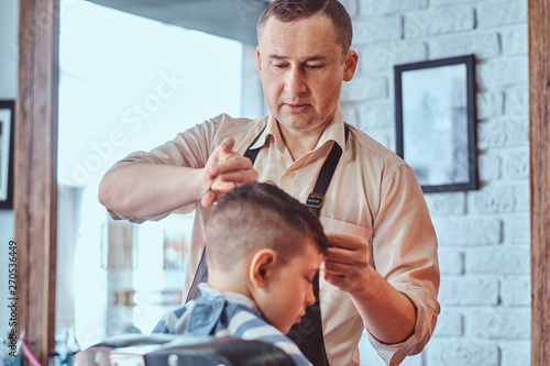 Attractive school boy is getting trendy haircut from mature hairdresser at fashionable hairdressing salon.