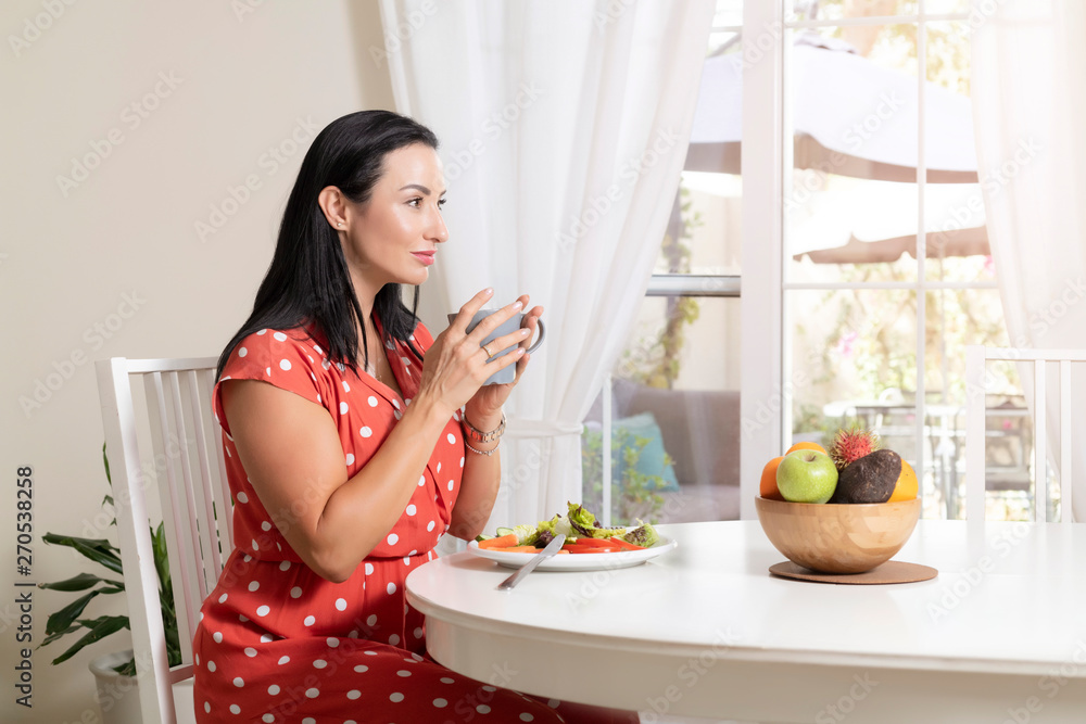 Beautiful brunette woman sitting wearing a red and white polka dot dress at dining table eating a healthy salad and drinking coffee or tea with a bowl of fruits on table with garden in the background