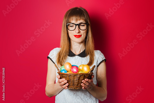 Young woman with a smile holds in her hands a basket of Easter eggs on a dark pink background. Concept of celebrating Easter, happy Easter