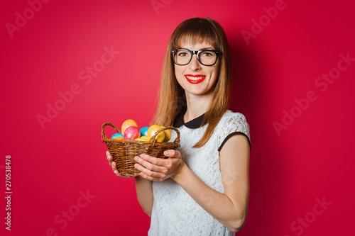 Young woman with a smile holds in her hands a basket of Easter eggs on a dark pink background. Concept of celebrating Easter  happy Easter
