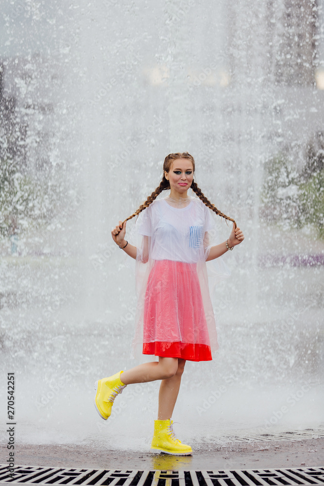 Young wet pretty girl with two braids in yellow boots and with transparent umbrella stands inside of fountain. Young wet pretty girl with two braids in yellow boots and with transparent umbrella stands inside of fountain.