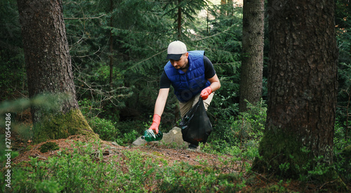 Adult man volunteer in red gloves removes plastic trash in forest, caring for environment.