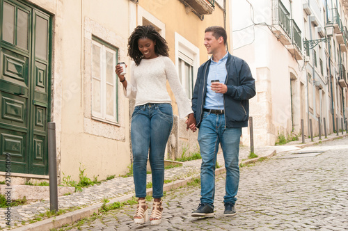 Happy Caucasian guy in love with her Afro American girlfriend. Mix raced couple holding hands, walking around old European city and drinking takeaway coffee. Dating concept