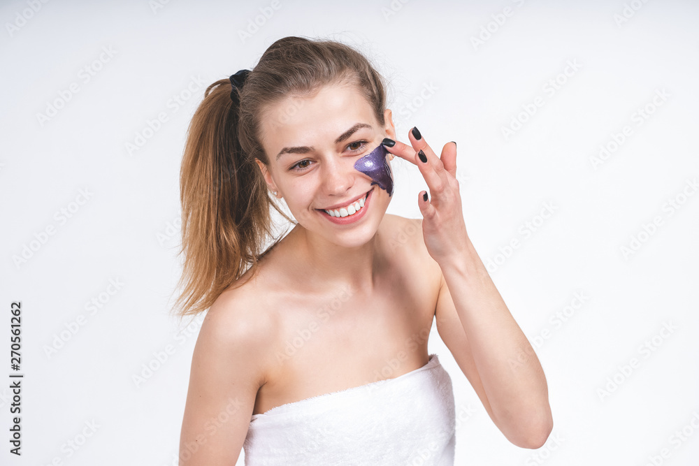 portrait young cheerful girl puts on face violet cosmetic mask. European, 22 years old, white, smiling, empty background, in a towel portrait young cheerful girl puts on face violet cosmetic mask. European, 22 years old, white, smiling, empty background, in a towel