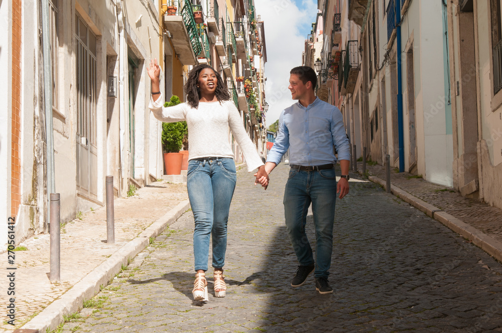 Positive cheerful multiethnic couple dating outdoors and chatting. African American girl and Caucasian guy holding hands and walking down old alley. Dating outdoors concept