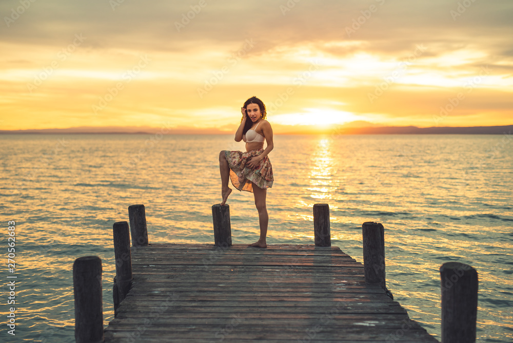 Happy woman enjoying the summery sunset at the lake on her vacation. The woman in bra and short summer skirt laughs and poses at the end of the wooden walkway Happy woman enjoying the summery sunset at the lake on her vacation. The woman in bra and short summer skirt laughs and poses at the end of the wooden walkway