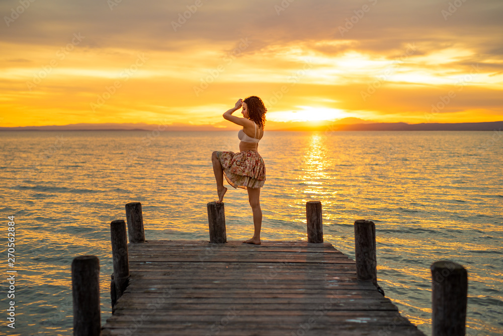 Happy woman enjoying the summery sunset at the lake on her vacation. The woman in bra and short summer skirt laughs and poses at the end of the wooden walkway
