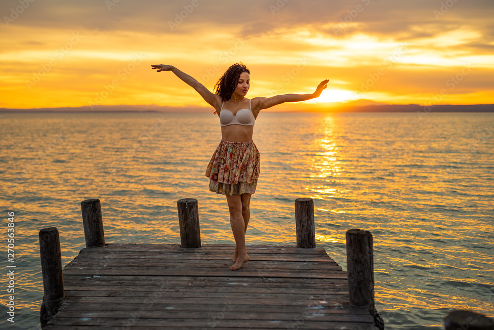Fototapeta premium Happy woman enjoying the summery sunset at the lake on her vacation. The woman in bra and a short summer skirt laughs and puts her arms in the direction of the sky