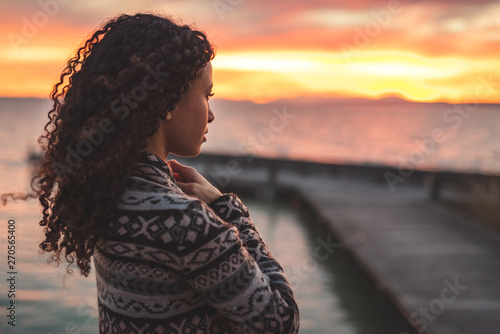 Thoughtful evening mood with a young Afro American woman standing on the promenade by the lake and looking towards the water and the setting sun. Burning sky photo