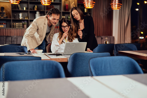 Charming buisnesswoman with brown hair and dressed elegant sitting at restaurant and looking at laptop. Next to her two colleagues standing and looking at laptop, too.