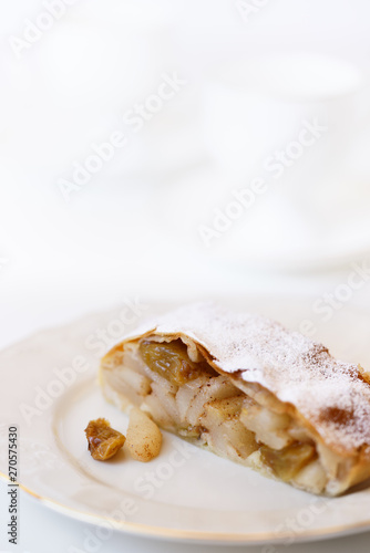 Apple strudel served on a white porcelain plate. Coffee cups in bokeh, white table, high resolution