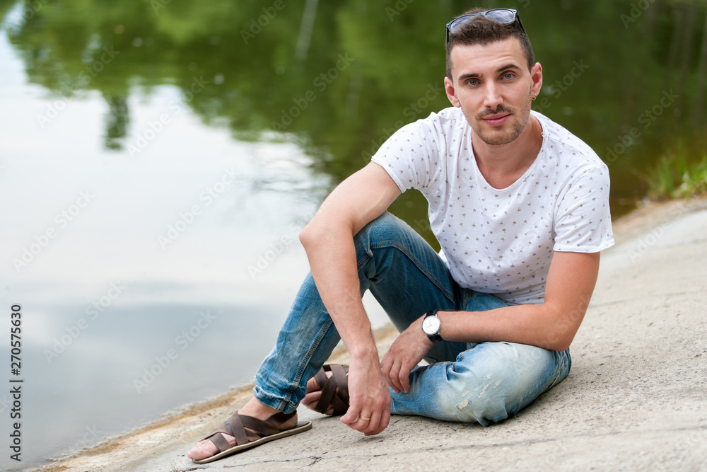 Young handsome man sitting next to lake. Water reflects trees.