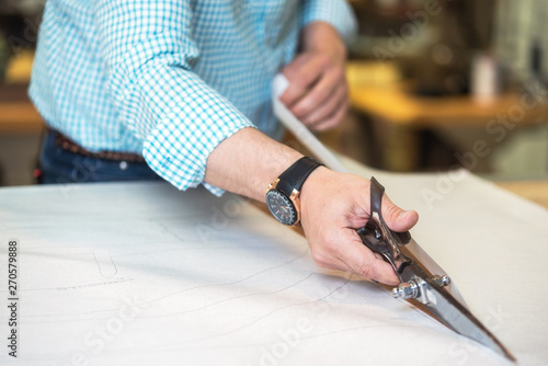 Tailor cutting out the marked pattern on fabric with large scissors on the workbench in his shop, close up view of his hands .