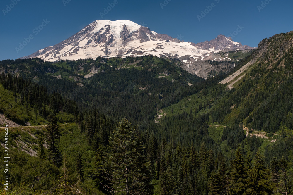 A snow capped mountain, Mount Rainier, at spring time with a forest of lush green pine trees in the foreground and road sweeping across the picture, nobody in the image