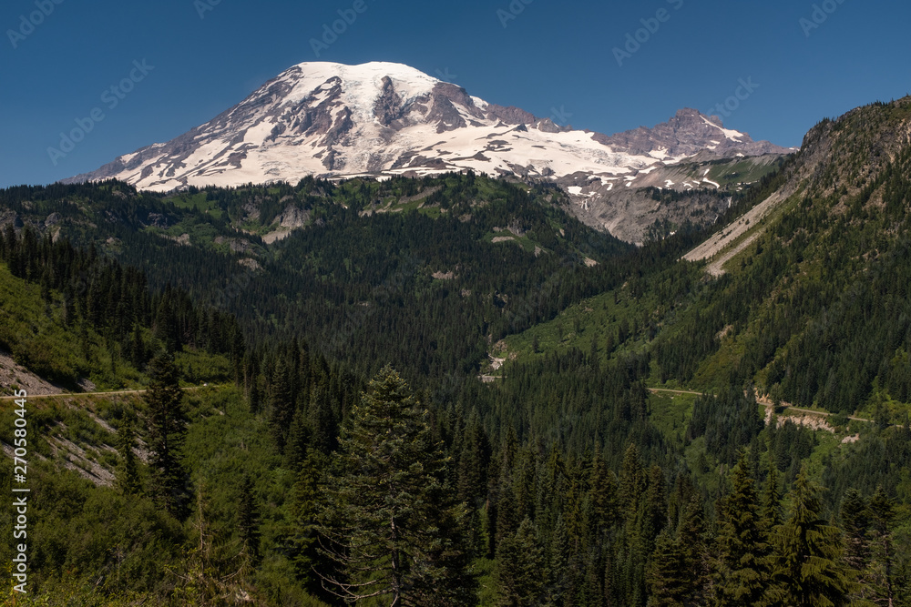 Fototapeta premium A snow capped mountain, Mount Rainier, at spring time with a forest of lush green pine trees in the foreground and road sweeping across the picture, nobody in the image