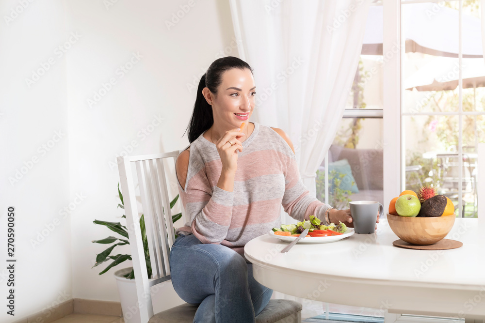 Beautiful brunette woman sitting at dining table eating a healthy salad and drinking coffee or tea with a bowl of fruits on table with garden in the background Beautiful brunette woman sitting at dining table eating a healthy salad and drinking coffee or tea with a bowl of fruits on table with garden in the background