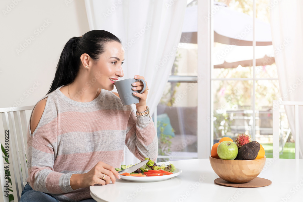 Beautiful brunette woman sitting at dining table eating a healthy salad and drinking coffee or tea with a bowl of fruits on table with garden in the background Beautiful brunette woman sitting at dining table eating a healthy salad and drinking coffee or tea with a bowl of fruits on table with garden in the background