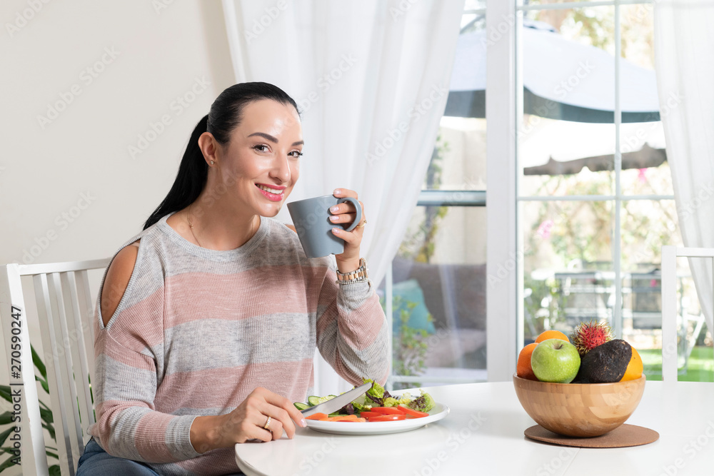 Beautiful brunette woman sitting at dining table eating a healthy salad and drinking coffee or tea with a bowl of fruits on table with garden in the background