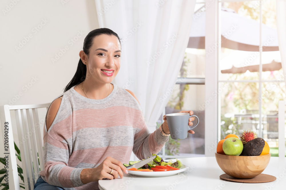 Beautiful brunette woman sitting at dining table eating a healthy salad and drinking coffee or tea with a bowl of fruits on table with garden in the background Beautiful brunette woman sitting at dining table eating a healthy salad and drinking coffee or tea with a bowl of fruits on table with garden in the background