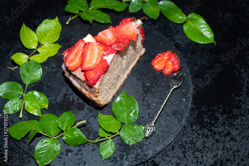 A piece of sponge cake with sour cream cream, fresh strawberries on top, lay on a black tray, around a spiral lined green rose leaves and fan-sliced ​​berries. 