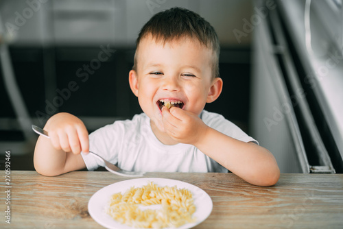 a little boy eats pasta in the form of a spiral in the afternoon in the kitchen on their own