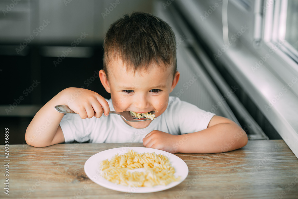 little boy eats pasta in the form of a spiral in the afternoon in the kitchen on their own