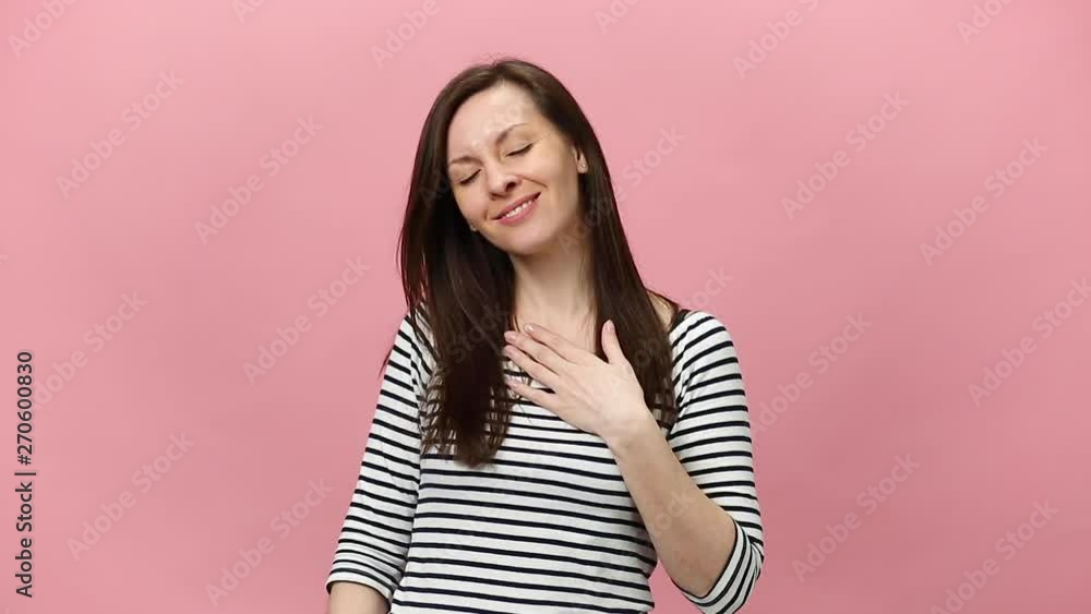Smiling mystery brunette young woman in striped shirt looking at camera with charming smile, sending air kiss isolated over pastel pink background in studio. People sincere emotions lifestyle concept. Smiling mystery brunette young woman in striped shirt looking at camera with charming smile, sending air kiss isolated over pastel pink background in studio. People sincere emotions lifestyle concept.