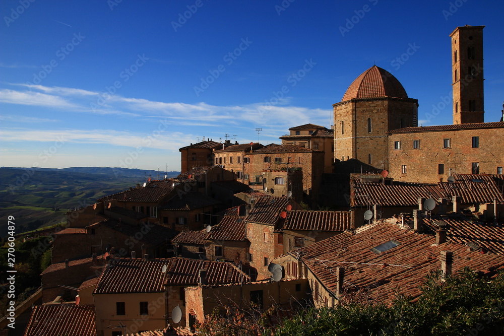 The roofs of the medieval town of Volterra overlook the autumn hills of Tuscany. Satellite dishes, medieval architecture and rustic tiles: epochs mix, just like blue and ochre.