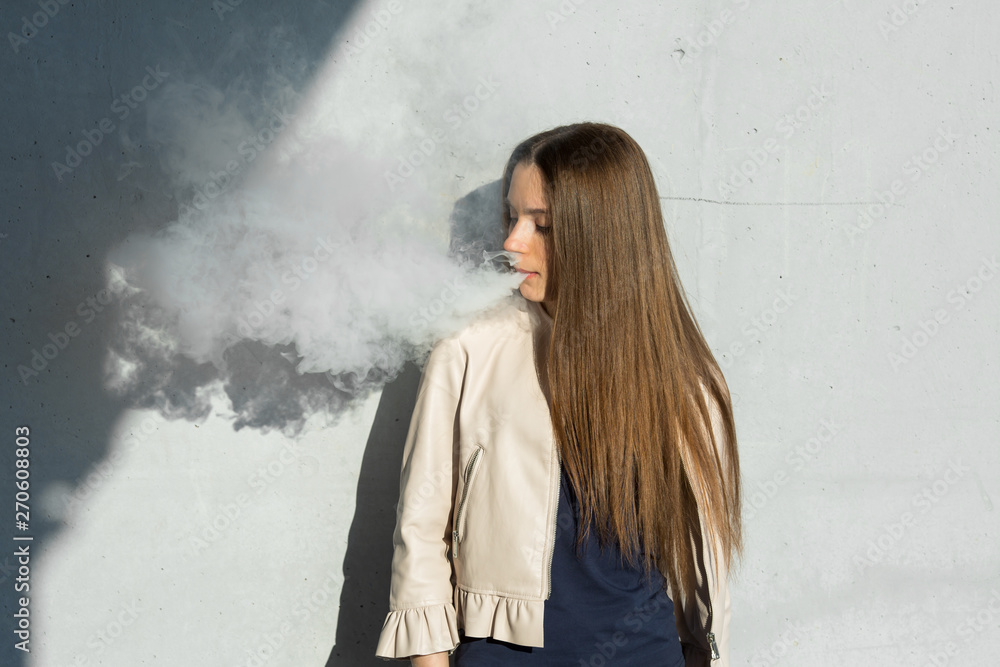 Vape teenager. Young cute girl in casual clothes smokes an electronic cigarette near the wall outdoors in summer day. Bad habit that is harmful to health. Vaping activity. Close up. Vape teenager. Young cute girl in casual clothes smokes an electronic cigarette near the wall outdoors in summer day. Bad habit that is harmful to health. Vaping activity. Close up.