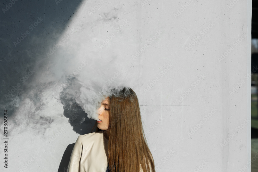 Vape teenager. Young cute girl in casual clothes smokes an electronic cigarette near the wall outdoors in summer day. Bad habit that is harmful to health. Vaping activity. Close up. Vape teenager. Young cute girl in casual clothes smokes an electronic cigarette near the wall outdoors in summer day. Bad habit that is harmful to health. Vaping activity. Close up.