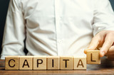 A man puts wooden blocks with the word Capital. The combination of material, intellectual and financial resources used for profit, additional material benefits. Concept of business and finance