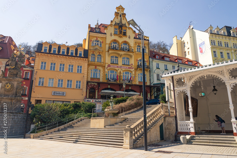 Outdoor sunny view of Holy trinity column, fountain and stairway beside Market colonnade, and background of Beautiful hotel's facade and colorful buildings in Karlovy Vary, Czech Republic..  
