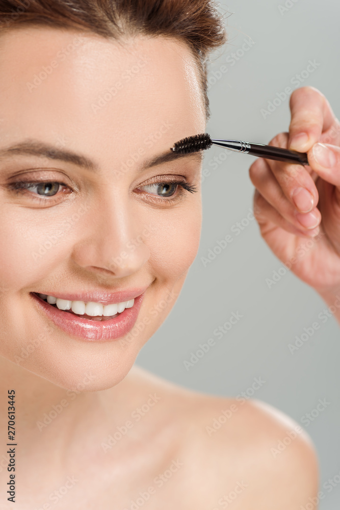 cropped view of makeup artist holding eyebrow brush near cheerful naked woman isolated on grey