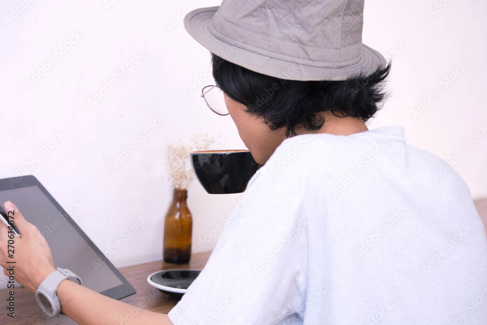 Back view. Young creative man in white shirt and padora hat sitting at table, using laptop while holding a cup of coffee Back view. Young creative man in white shirt and padora hat sitting at table, using laptop while holding a cup of coffee
