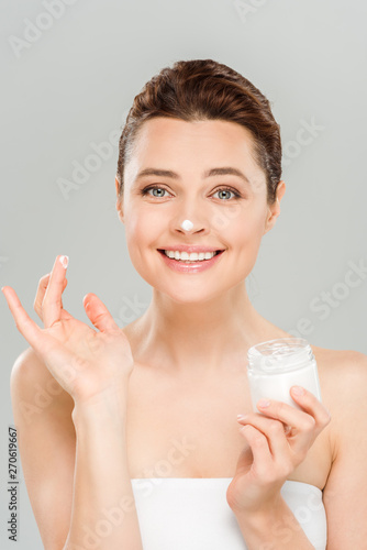 cheerful woman applying face cream and smiling isolated on grey